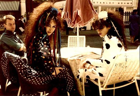 Jill in dark outfit with coloured polka dots and rose in white with huge black polka dots sit at an outdorr cafe table in blazing sunshine, turning towards camera. Manager David Balfe sits at the far side of the table with arms folded.