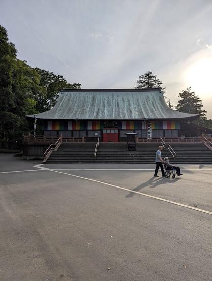 A man pushing a woman in a wheelchair by a big plaza. A big one-floor temple hall is in the background of the photo. The late afternoon sun shines bright in the top right of the photo, making nice diagonal and long shadows out of the two people.