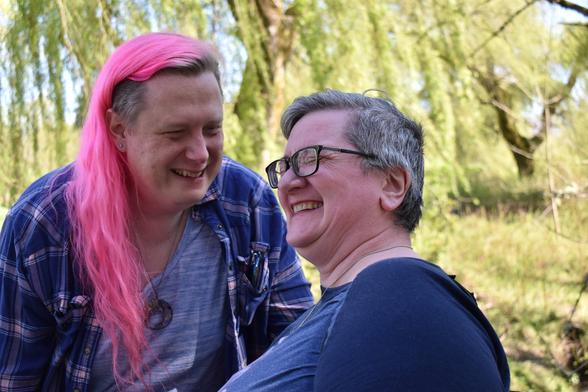 Two people, one with long pink hair wearing a flannel and the other with short gray hair and glasses, standing in front of willow trees laughing together.