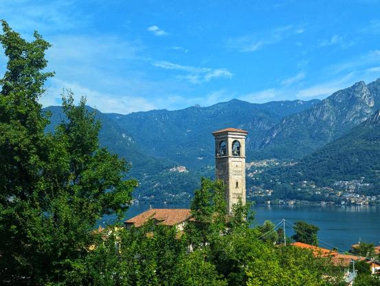 A scenic, slightly high-angle shot captures a stone bell tower with arched openings at its top, housing visible bells. The tower stands amidst lush green trees and terracotta-roofed buildings, suggesting a village or town setting. Behind the buildings, a tranquil blue lake stretches horizontally across the frame, reflecting the clear, bright blue sky dotted with wispy white clouds. In the distance, majestic, dark green mountains rise against the sky, completing the picturesque landscape. The overall impression is one of a peaceful and idyllic European countryside scene.