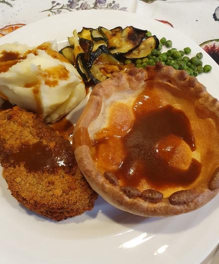 A plate of food featuring mashed potatoes with gravy, a breaded veggie cutlet, roasted courgette slices, green peas, and a Yorkshire pudding filled with gravy.