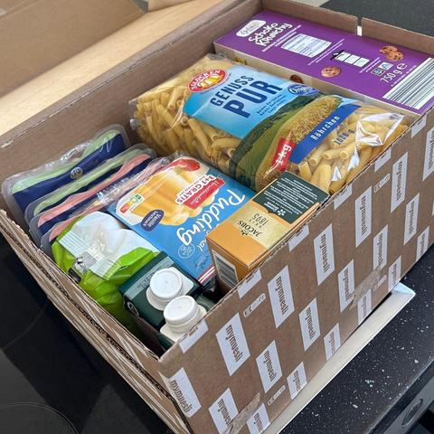 Das Bild zeigt eine offene Kartonbox auf einer Küchentheke, gefüllt mit verschiedenen Lebensmitteln. Im Vordergrund sind mehrere Packungen Pasta zu sehen, darunter eine große Packung "Genius Pur" Röhrchenpasta und eine Packung "Schule Kräuter" Cookies. Daneben befinden sich mehrere Packungen von "Pudl" Joghurt in verschiedenen Geschmacksrichtungen, wie zum Beispiel "Strawberry" und "Vanille". Es gibt auch mehrere Trinkdosen, darunter eine grüne Packung, die wahrscheinlich Milch oder ein anderer Getränk enthält, sowie eine Packung von "Jacobs" Bohnen. Die Kartonbox ist mit dem Logo "mymuesli" versehen. Die Lebensmittel sind ordentlich in der Box angeordnet, was auf eine sorgfältige Auswahl hinweist.