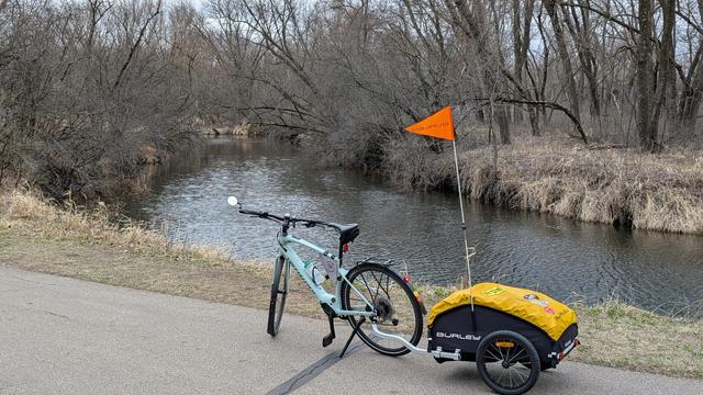 Sage green Vado SL ebike with yellow tarped Burley Nomad trailer with orange pennant flag parked on the Bear Creek Trail with the creek behind, surrounded by mostly bare trees.