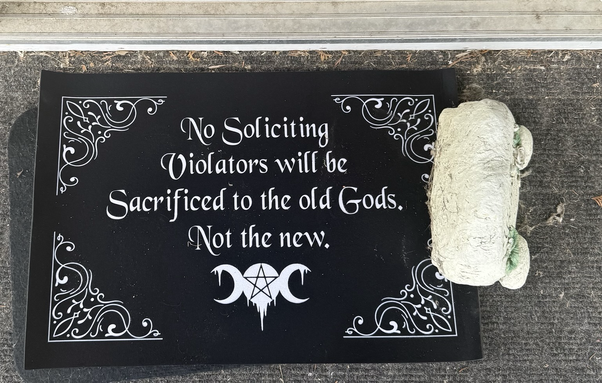 Black doormat with triple moon and star symbol on bottom, and scrollwork on the sides. White script reads ; no soliciting , violators will be sacrificed to the old  Gods not the new.