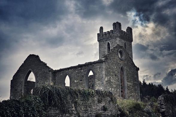A photo of the weathered stone ruins of Castlemacadam Church in Ireland under a dramatic, cloudy sky. Ivy grows on the lower walls.