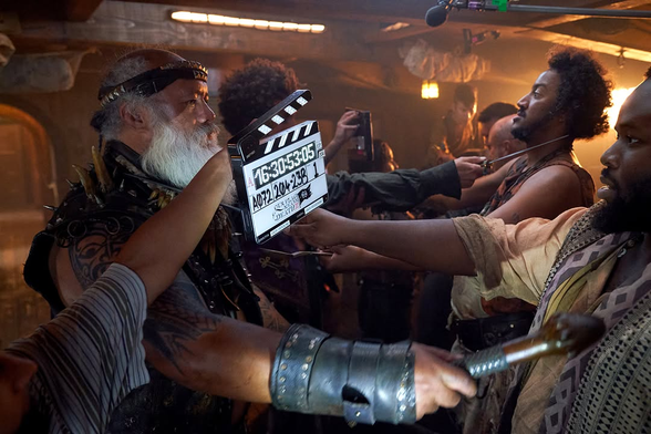 Behind-the-scenes photo of a scene in Season 2, Episode 4: Stede's and Blackbeard's crews stand facing each other, holding weapons to each other's throats. A clapperboard is being held up between them and the camera. David Fane (Fang), Samson Kayo (Oluwande) and Samba Schutte (Roach) are at the front, with the rest of the cast in the background. They are all in position, their postures stiff as they wait for “action” to be called.
