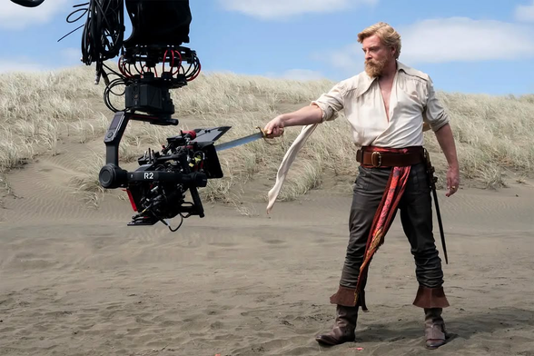 BTS photo from the OFMD Season 2 opening scene: Rhys Darby as Stede Bonnet, seen from the side, stands on a beach in his campy swashbuckler outfit and a full beard, one arm outstretched to point a sword at a camera in front of him, which is mounted at about hip level on a robotic camera arm.