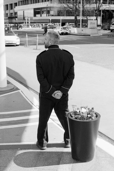 A doorman stands at attention with hands folded behind before a blooming flower pot.