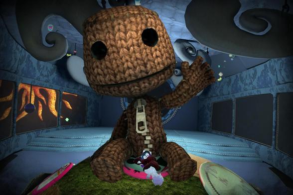 The image shows a large, knitted, sack-like creature, similar to the character Sackboy from the LittleBigPlanet video game series. It is sitting in a room with a blue, patterned interior. Two smaller characters are lying on a pink cushion at its feet. The overall scene appears to be from a video game.