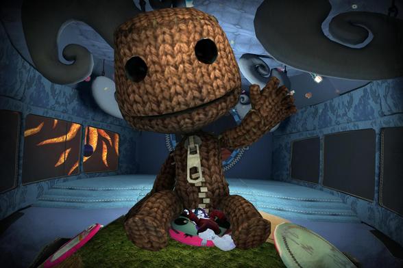 The image shows a large, knitted, sack-like creature, similar to the character Sackboy from the LittleBigPlanet video game series. It is sitting in a room with a blue, patterned interior. Two smaller characters are lying on a pink cushion at its feet. The overall scene appears to be from a video game.