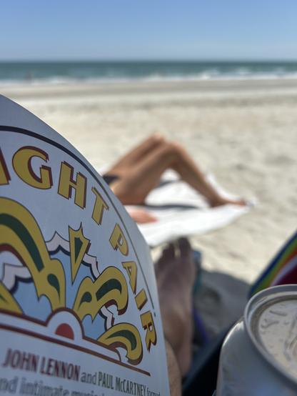 A photo of the beach from my perspective. A magazine page. The top of a beer can. The legs of somebody getting a tan. The sand of the beach and the sea. Blue sky and sun.