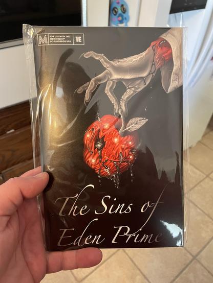 A hand with gray skin holds a red, stylized apple that drips with liquid. The apple features intricate designs and details. The title "The Sins of Eden Prime" is displayed prominently at the bottom.