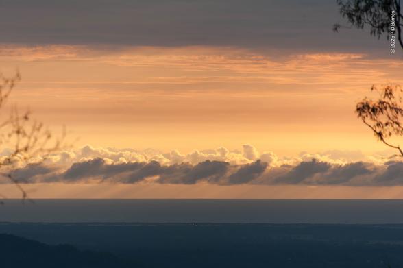 High view at sunrise, looking out over dark plains to the sea and horizon. Above the horizon is a band of cumulus with regular, repeating shapes, those at the front still dark and those behind lit by the rising sun. The whole sky is a peach colour, contrasting the two dark bands of land and sea, and a further band of dark cloud at the top of the image. Overall the image looks like an abstract Art Nouveau frieze in peach.