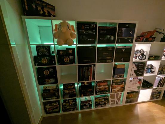 Left Kallax with green hue coming from behind the shelf. Compartments are mostly filled with boardgames, plus one mastodon plushy
