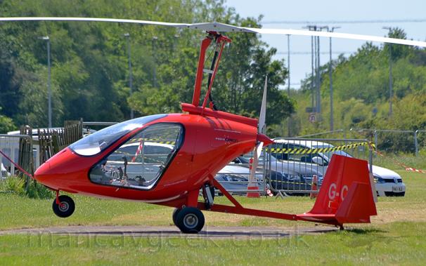 Side view of a bright red, 2 seat autogyro, parked facing to the left.
The aircraft has a sleek red body, with large, fully glazed doors under a large white rotor, with a smaller white pusher propellor at the top of the rear fuselage.
A low, thin red tail boom carries the red tail, a large central fin with smaller end-plates on the horizontal stabiliser.
The first couple of letters of the registration is visible on the central fin in grey.
There are small, grey and black "M24 Orion" titles on the top of the fuselage, just ahead of the pusher prop.
Green grass fills the foreground, with several grey cars lined-up side-by-side behind a grey metal fence in the background, with trees in the distance under hazy grey skies.