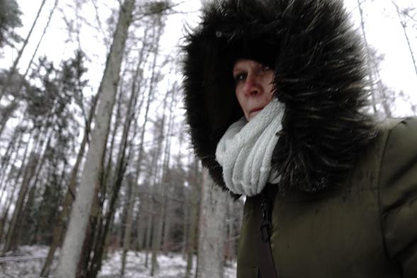 The image shows a person in a winter setting, wearing a thick, fur-lined hooded coat and a scarf. The background consists of tall trees in a snowy forest.