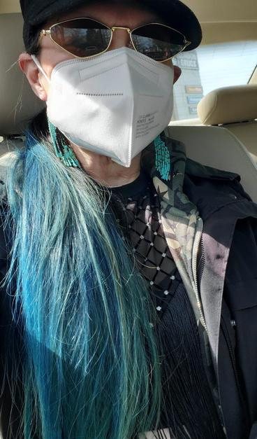 Me, wearing a white KN95 protective mask that covers nose and mouth. Wearing gold-rimmed sunglasses and a black hat. With long, blue hair and distinctive turquoise beaded black raven earrings. Wearing a black jacket over a camo hoodie and black top with a pattern of black fringe and diamonds. The car's interior is visible, with a light-colored leather seat. Sunlight fills the interior of the car creating sunny conditions.