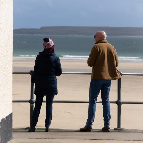 A man and woman stand talking by the sea. The woman looks out to sea whilst the man turns to her. Immediately in front of them is a metal railing. The sand on the beach is golden and white waves break in an aquamarine sea.