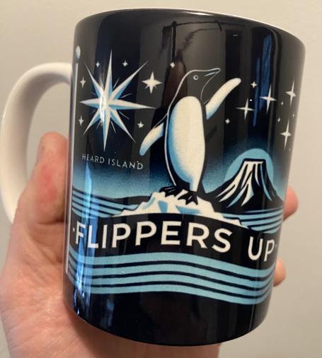A picture of a coffee mug from the Heard Island  Government store. It features a penguin with its flipper up standing atop a motto that says FLIPPERS UP.