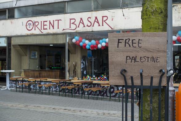 color photograph, landscape format. in the background empty shop windows with white-red-blue balloons, above them the inscription “orient bazaar”. on the right, wedged into a barrier, a piece of wood with the inscription “free palestine”.