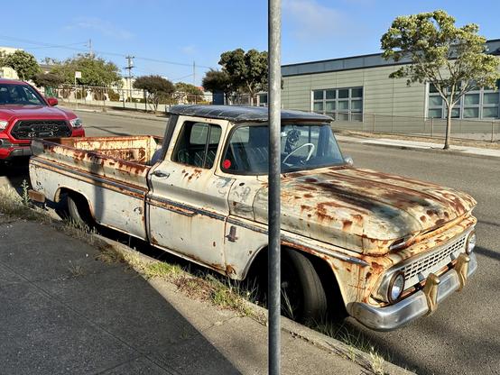 Photograph from a front three-quarters view of a vintage Chevrolet pickup. At one time, vehicle was painted a creamy white with a contrasting black top to the passenger compartment but extensive rust, corrosion, and sun-bleaching show the truck’s age. The truck is parked on a low-density urban street, with trees and a school visible in the background against a blue sky.