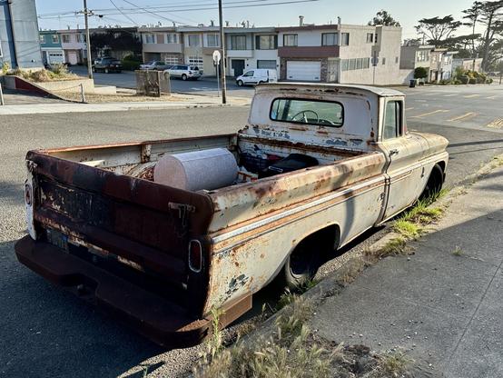 Photograph from a rear three-quarters view of a vintage Chevrolet pickup. At one time, vehicle was painted a creamy white, but extensive rust, corrosion, and sun-bleaching show the truck’s age. The truck’s tailgate is dark in shadow, while its cargo bed holds a cylindrical tank of some kind. The vehicle is parked on a low-density urban street, with trees and several houses visible in the background.