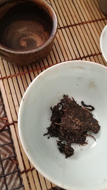 A dark and stained tea cup and a pale gaiwan sit on a bamboo mat. There is a little pile of as-yet un-steeped puer leaves in the gaiwan.