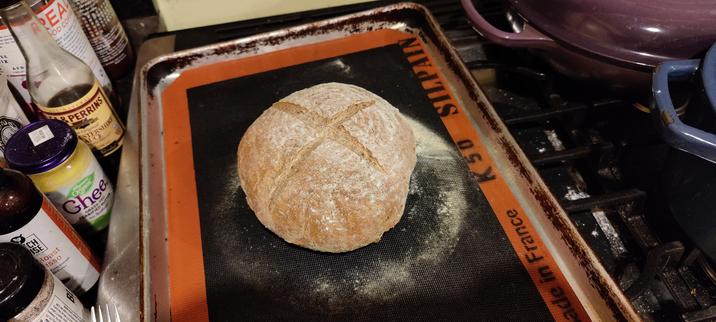 A round loaf of rye bread with an expansion cross cut across the top, sitting on a black baking mat in a half- sheet pan.
