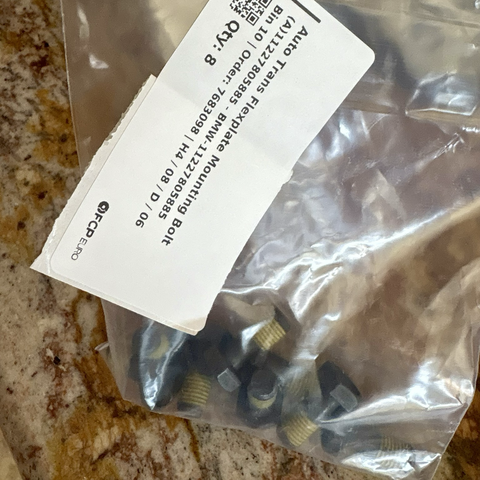 A clear plastic bag containing dull bronze-colored bolts sits on my brown granite kitchen countertop. A label on the bag identifies the contents as "Auto Trans Flexplate Mounting Bolts."