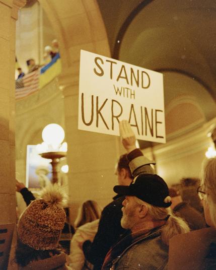 A photograph of the Minnesota State Capitol building, softly lit in warm tones. A banner reading “Stand with Ukraine” hangs prominently on the balcony, accompanied by the Ukrainian and American flags. The architecture frames the message, highlighting solidarity in a quiet, powerful way. Captured on Fuji 200 film with a Canon Elan camera.
