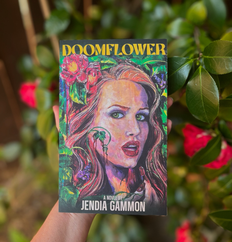 Doomflower by Jendia Gammon, held in front of red camellia flowers by the author. The main character’s name is Camellia Dume, and she can be seen on the cover with long red hair, green eyes, red lipstick and nails, surrounded by camellia flowers and kudzu blooms and leaves, with blood dripping from them.