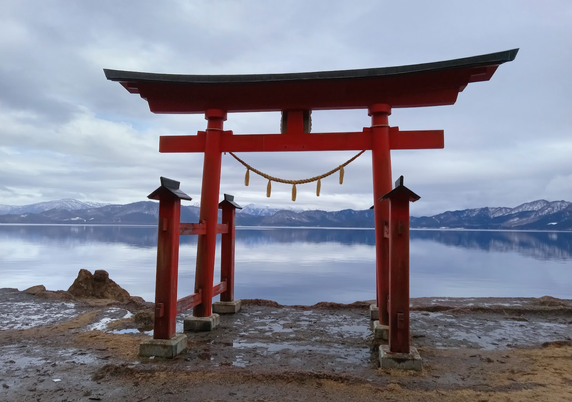 photo of a red torii gate on the shore of Lake Tazawa in Akita, Japan ... the lake can be seen behind the gate with mountains in the distance ... the ground is muddy with puddles all around