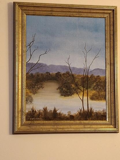 Within a gold frame  a painting of a body of water reflecting the sky  with hills beyond.