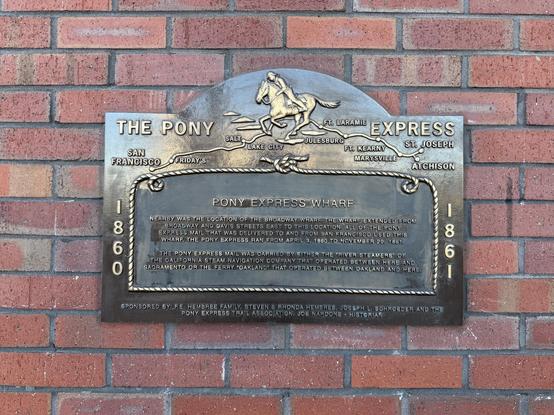 Photograph of a historical marker affixed to a brick wall. The upper portion of the plaque depicts a map of the Pony Express route from St. Joseph, Missouri, west to San Francisco. It is surmounted by a galloping horse and rider. Below it, a stylized lasso surrounds text that reads:

“PONY EXPRESS WHARF

NEARBY WAS THE LOCATION OF THE BROADWAY WHARF. THE WHARF EXTENDED FROM BROADWAY AND DAVIS STREETS EAST TO THIS LOCATION. ALL OF THE PONY EXPRESS MAIL THAT WAS DELIVERED TO AND FROM SAN FRANCISCO USED THIS WHARF. THE PONY EXPRESS RAN FROM APRIL 3, 1860 TO NOVEMBER 20, 1861.
THE PONY EXPRESS MAIL WAS CARRIED BY EITHER THE ‘RIVER STEAMERS’ OF THE CALIFORNIA STEAM NAVIGATION COMPANY THAT OPERATED BETWEEN HERE AND SACRAMENTO OR THE FERRY ‘OAKLAND’ THAT OPERATED BETWEEN OAKLAND AND HERE.”

This text, and the lasso, are together bracketed by the dates 1860 and 1861.