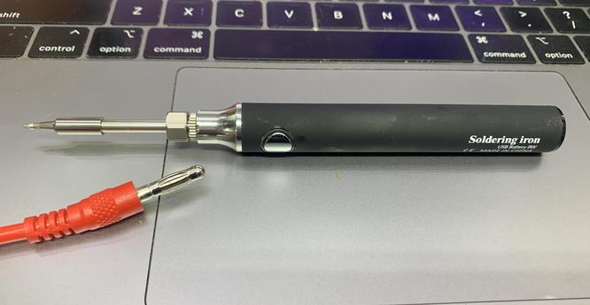A petite cordless soldering iron sits on the deck of a macbook.   It's handle is about the size of 2xAA cells, it has a silver button, no screen and a replaceable tip.