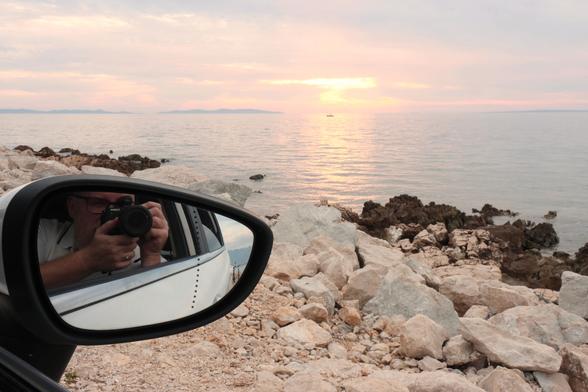 In primo piano lo specchietto di un'automobile in cui si riflette il fotografo che impugna una macchina fotografica, sullo sfondo il mare al tramonto.