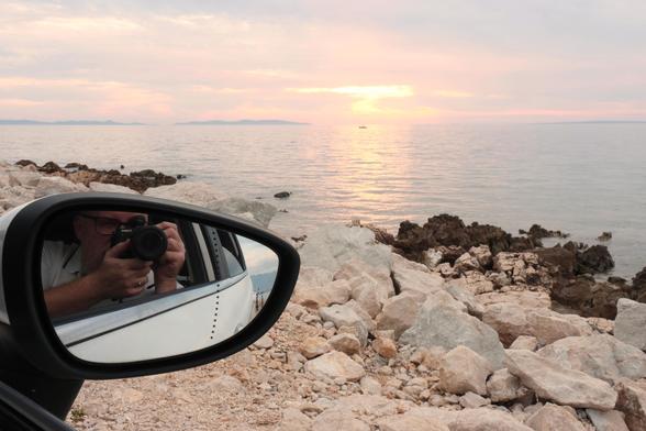 In primo piano lo specchietto di un'automobile in cui si riflette il fotografo che impugna una macchina fotografica, sullo sfondo il mare al tramonto.