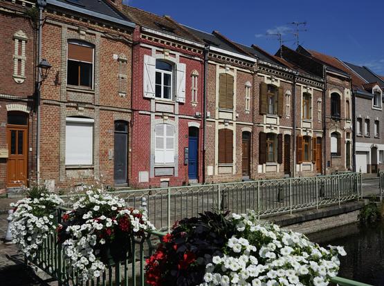 Vue de biais sur un alignement de vieilles maisons en briques à deux niveaux le long d’une voie longeant un canal. Les rambardes du canal sont fleuries.
