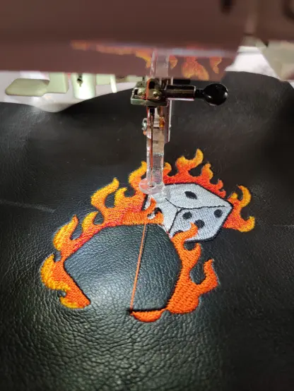 Man sieht schwarzes Kunstleder, das in einer Stickmaschine liegt. Große Teile des Motives sind schon fertig: 2 sechseitige weiße Würfel, die von Flammen umgeben sind, die in einem Farbverlauf rot nach gelb ausgeführt sind. Ein Würfel fehlt dabei noch komplett.