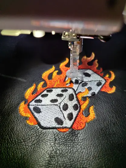 Man sieht schwarzes Kunstleder, das in einer Stickmaschine liegt. Das Motiv ist hier nahezu fertig: 2 sechseitige weiße Würfel, die von Flammen umgeben sind, die in einem Farbverlauf rot nach gelb ausgeführt sind.