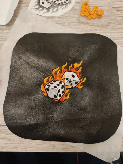 Das fertig bestickte schwarze Kunstleder liegt nun auf dem Tisch. Es ist noch nicht zugeschnitten, aber das Motiv ist fertig: 2 sechseitige weiße Würfel, die von Flammen umgeben sind, die in einem Farbverlauf rot nach gelb ausgeführt sind. 
Auf dem Tisch sieht man zum Teil das Ursprungsmotiv auf Papier ausgedruckt sowie eine kleine Schachtel mit orangen Druckknöpfen.
