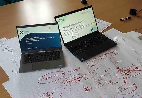Das Bild zeigt zwei offene Laptops auf einem Tisch, der mit Papier und Notizen bedeckt ist. Der Laptop links zeigt eine Präsentationsfolie mit dem Titel "SKOS in der Praxis" und dem Logo von NFDI for Objects. Der Laptop rechts zeigt eine Folie mit dem Titel "Objekt? Nebensache? Hauptsache? Ereignis!" und dem Logo des SODA-Projekts. Auf dem Papier sind verschiedene Notizen und Diagramme in roter Tinte zu sehen. Ein Marker liegt auf dem Tisch, und im Hintergrund sind weitere Papierstapel und ein Computermauszeiger zu erkennen.