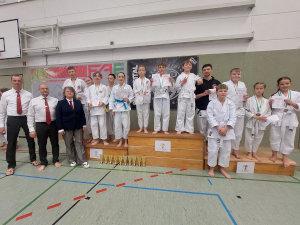 Gruppenbild mit allen Teilnehmern des ersten Kaminari-Arashi-Cups auf dem Siegerpodest

Rechts neben dem Podest stehen drei Kampfrichter und Trainer des Dojos Tameshiwari.