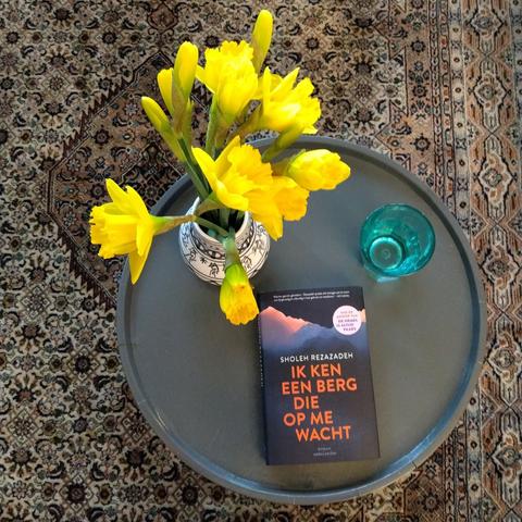 Foto van narcissen met een boek van Sholeh Rezazadeh op een ronde tafel van bovenaf genomen. De titel: Ik ken een berg die op me wacht.
