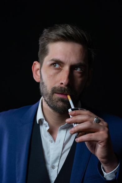 A person with short, styled hair, wearing a white shirt, a vest, and a blue suit jacket, holds a cigarette at their lips while gazing off to the side. A silver ring on one finger is visible as they raise the cigarette box. The black background provides strong contrast.