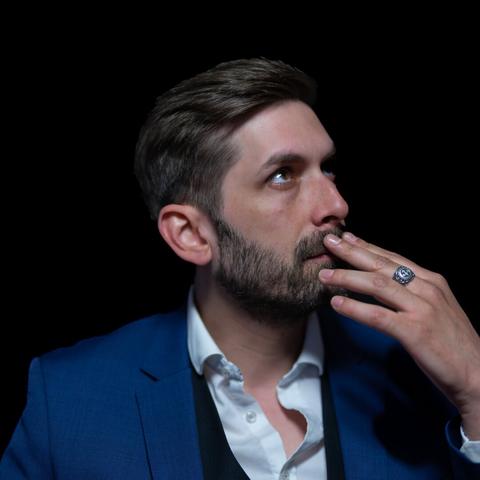 A head-and-shoulders portrait of a person with short, styled hair, wearing a white shirt, a vest, and a blue suit jacket. They glance upward with fingertips gently touching their lips, and a silver ring catches the light. The black background and subtle lighting draw attention to the subject’s contemplative gaze.
