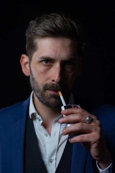 A person with short, styled hair, wearing a white shirt, a vest, and a blue suit jacket, holds a cigarette at their lips, looking directly at the camera. The black background and focused lighting emphasise their intense gaze and facial features. One hand grips the cigarette box, revealing a silver ring on their middle finger.