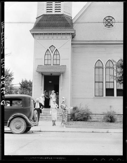 A black and white photograph depicting a scene outside of a church, likely taken during the early to mid-20th century. The building features Gothic architectural elements such as pointed arch windows and a steeple with a weather vane at its peak.

In front of the church stands an old-fashioned sedan parked on a street adjacent to a paved sidewalk where several people are congregating near steps leading into the church entrance. There's a woman, possibly in her 30s or early 40s wearing a flowing white dress with floral patterns and another older woman beside her; both appear happy.

Two men in suits stand by the car—one smoking—and two women also dressed in light-colored dresses are seen on the sidewalk. The attire of everyone is indicative of mid-20th-century fashion, suggesting this might be Sunday morning or a special event like church service attendance.

Various vegetation and shrubs adorn what looks to be an urban environment with paved roads indicating it's not rural. A watermark text overlay reads "Dorothea Lange - National Photo Company," which indicates the photographer who captured this moment in history as well as associated archives for additional information about her work during that era, including coverage of important events such as The Scopes Trial held in Dayton, Tennessee—a town famous for its courtroom drama involving evolution debates.