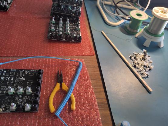 Picture of hardware making setup. You can see red bubble wrap on a wooden table with black circuit boards in various states, next to yellow pliers under blue spring cord. Next to that on a blue covering is a pile of components next to a singular chopstick and two rolls of soldering tin.