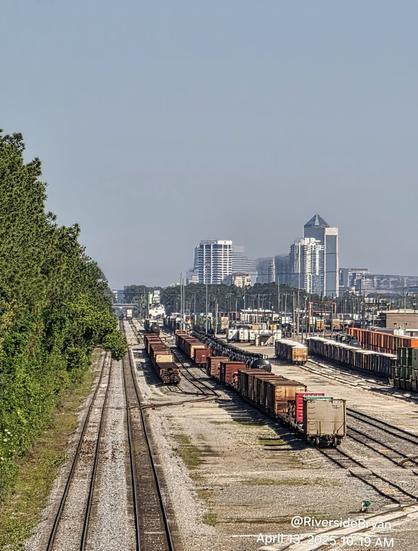 Overlooking a railyard with many different track lines, some with colorful rail cars lined up. In the background the downtown skyline towers above.